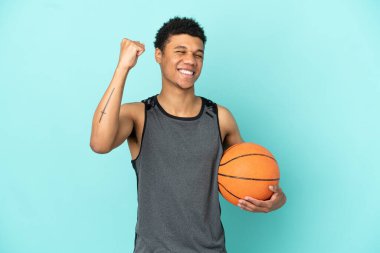 Basketbolcu Afrikalı Amerikalı adam mavi arka planda izole edilmiş zaferi kutluyor.