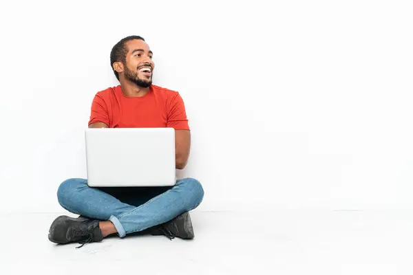 年轻的厄瓜多尔男人 拿着笔记本电脑 坐在地板上 被白色的背景隔离着 开心地微笑着 — 图库照片