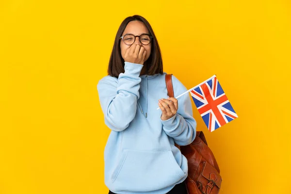 Young latin woman holding an United Kingdom flag isolated on yellow background happy and smiling covering mouth with hands