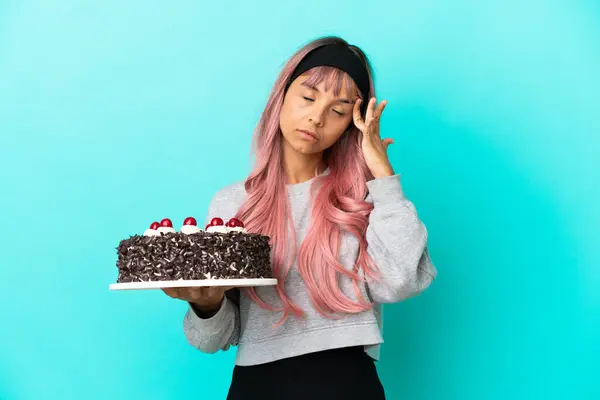 Young woman with pink hair holding birthday cake isolated on blue background with headache