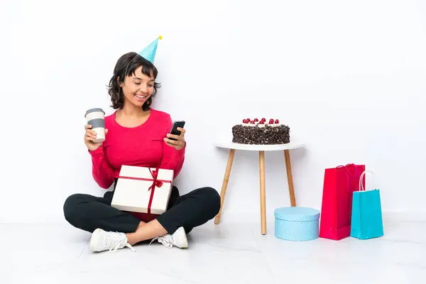 Young girl celebrating his birthday sitting on the floor isolated on white background holding coffee to take away and a mobile