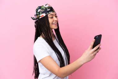 Örgülü genç kız izole edilmiş pembe arka planda cep telefonuyla selfie çekiyor.