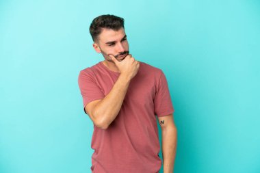 Kafkasyalı genç adam mavi arka planda izole edilmiş. Şüpheleri var ve kafası karışmış bir yüz ifadesi var.