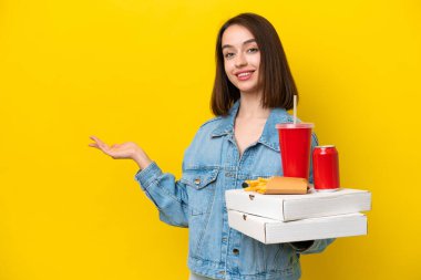 Sarı arka planda izole edilmiş fast food tutan genç Ukraynalı kadın davet edilmek için ellerini yana uzatıyor.