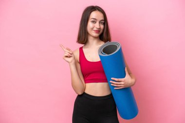 Genç sporcu Ukraynalı kadın yoga derslerine gidiyor. Elinde pembe arka planda izole edilmiş bir minder var. Ürünü sunmak için yan tarafa bakıyor.
