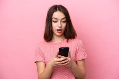 Pembe arka planda izole edilmiş genç Ukraynalı kadın kameraya bakarken şaşırmış bir ifadeyle cep telefonunu kullanıyor.
