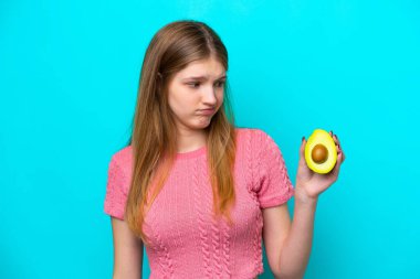 Teenager Russian girl holding an avocado isolated on blue background with sad expression