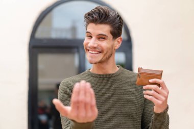 Açık havada cüzdan tutan genç beyaz bir adam, elini kolunu sallayarak gelmesi için davet ediyor. Gelmene sevindim.