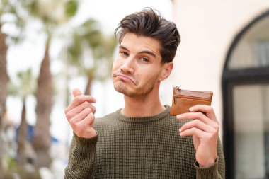 Genç, beyaz bir adam elinde cüzdan ve üzgün bir ifadeyle dışarıda bekliyor.