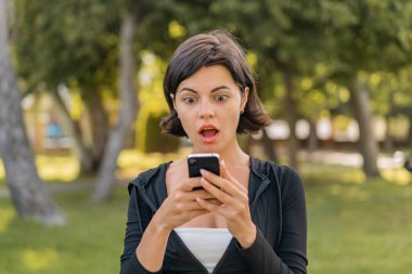 Genç, güzel bir Bulgar kadın açık havada kameraya bakarken şaşırmış bir ifadeyle cep telefonunu kullanıyor.
