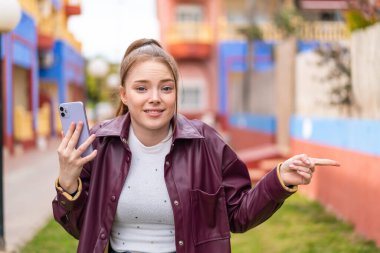 Açık havada cep telefonu kullanan genç ve güzel bir kız şaşırmış ve işaret ediyor.