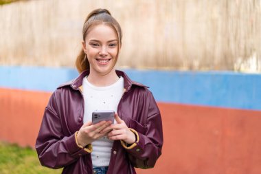 Genç ve güzel bir kız dışarıda cep telefonu kullanıyor. Omuzlarını kaldırırken şüpheli hareketler yapıyor.