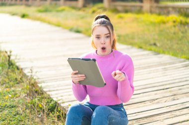 Açık havada elinde tablet tutan genç ve güzel bir kız şaşırmış ve ön tarafı işaret ediyor.