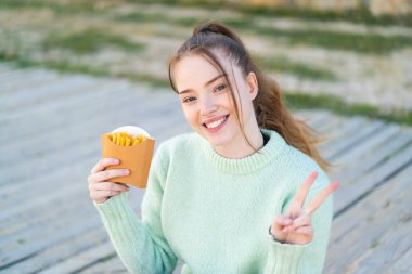 Genç ve güzel bir kız elinde cipslerle dışarıda gülümsüyor ve zafer işareti gösteriyor.