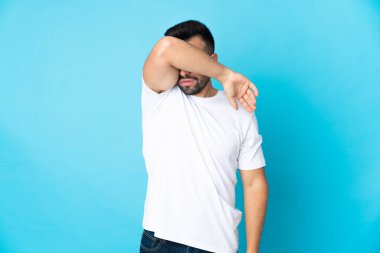 İzole edilmiş mavi arka planda beyaz bir adam elleriyle gözlerini kapatıyor.