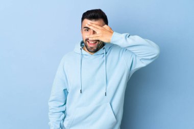 İzole edilmiş mavi arka plan üzerinde beyaz bir adam. Gözleri elleriyle kaplıyor ve gülümsüyor.