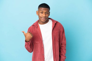 Mavi arka planda izole edilmiş genç latin adam bir ürün sunmak için kenarı işaret ediyor
