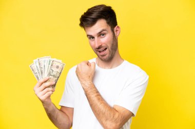 Young caucasian man taking a lot of money isolated on yellow background celebrating a victory