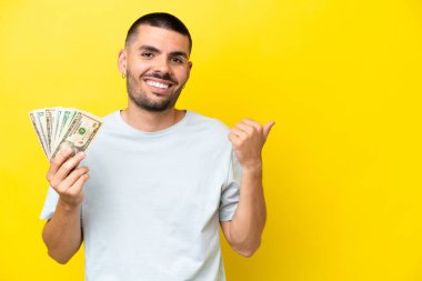 Young caucasian man taking a lot of money isolated on yellow background pointing to the side to present a product