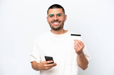 Beyaz arka planda izole edilmiş genç beyaz bir adam cep telefonuyla kredi kartıyla alışveriş yapıyor.