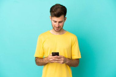 Mavi arka planda izole genç beyaz adam cep telefonuyla bir mesaj gönderiyor.