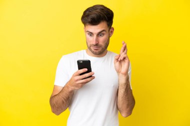 Beyaz tenli yakışıklı adam sarı arka planda izole edilmiş ve parmakları çapraz cep telefonu kullanıyor.