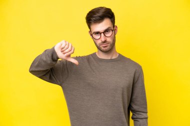 Sarı arka planda izole edilmiş genç beyaz adam olumsuz ifadelerle baş parmağını aşağı gösteriyor.