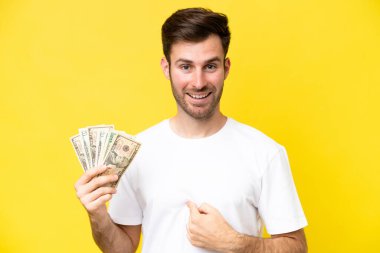 Kafkasyalı genç bir adam, sarı arka planda, şaşırtıcı bir yüz ifadesiyle izole edilmiş bir sürü para alıyor.