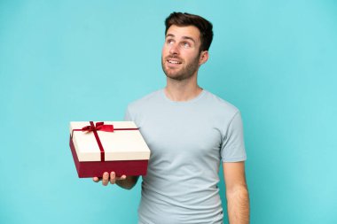 Elinde mavi arka planda izole edilmiş bir hediye tutan beyaz bir adam gülümserken yukarı bakıyor.