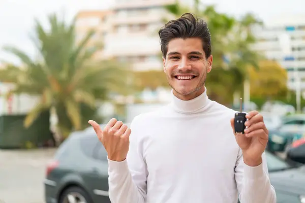 Stock image Young caucasian man holding car keys at outdoors pointing to the side to present a product