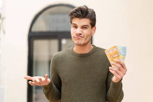 Young Caucasian Man Taking Lot Money Outdoors Making Doubts Gesture — Stock Photo, Image
