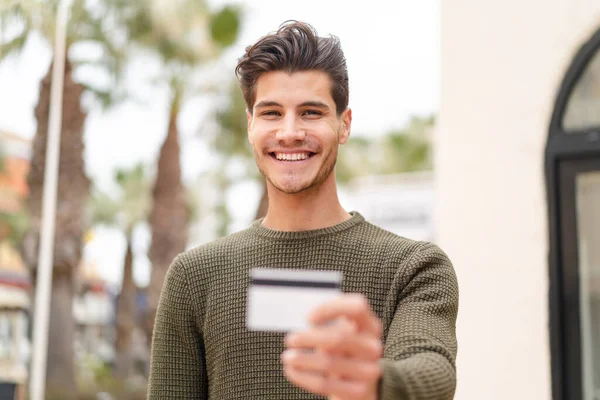 Açık havada genç beyaz bir adam elinde mutlu bir ifadeyle bir kredi kartı tutuyor.