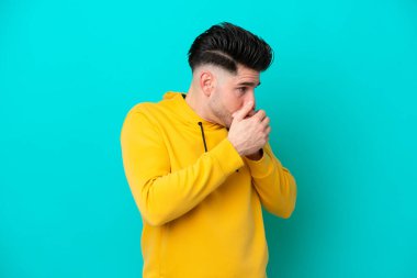 Genç beyaz adam mavi arka planda izole edilmiş ağzını kapatıyor ve yan tarafa bakıyor.