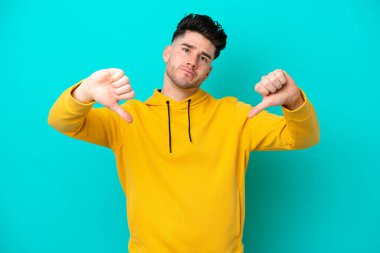 Mavi arka planda izole edilmiş genç beyaz bir adam baş parmağını iki eliyle aşağı gösteriyor.