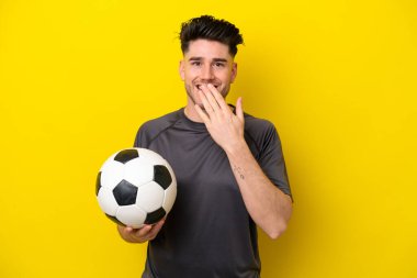 Yakışıklı genç futbolcu sarı arka planda izole edilmiş mutlu ve gülümseyen elleri ile ağzını kapatıyor.
