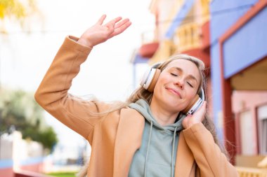 Genç sarışın kadın açık havada müzik dinliyor ve dans ediyor.