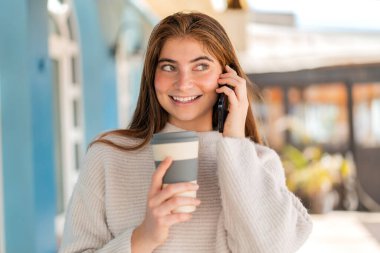 Genç beyaz bir kadın cep telefonu kullanıyor ve elinde mutlu bir ifadeyle kahve tutuyor.
