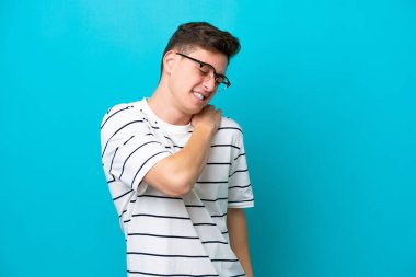 Genç ve yakışıklı Brezilyalı bir adam mavi arka planda izole edilmiş, omuz ağrısı çekiyor.