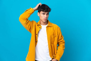 Soyutlanmış mavi arka planı olan genç bir adam kafasını kaşırken şüpheye düşüyor.