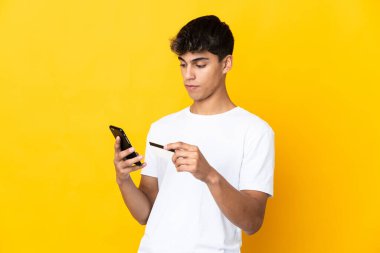 Young man over isolated yellow background buying with the mobile with a credit card