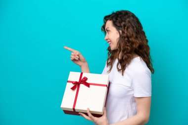 Elinde mavi arka planda izole edilmiş bir hediye tutan beyaz bir kadın bir ürünü sunmak için yan tarafı işaret ediyor.