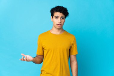 Genç Venezuelalı adam mavi arka planda izole edilmiş, şüphe uyandıran hareketler yapıyor.