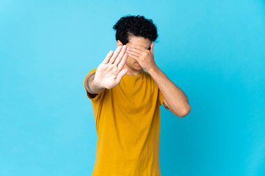 Genç Venezuelalı bir adam mavi arka planda izole edilmiş. Durma hareketi yapıyor ve yüzünü kapatıyor.