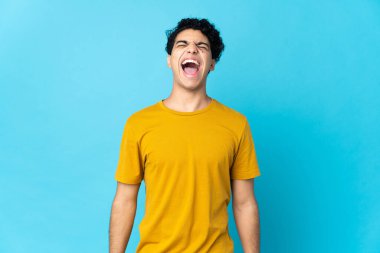 Genç Venezuelalı bir adam mavi arka planda izole edilmiş ve ağzı açık bağırıyor.