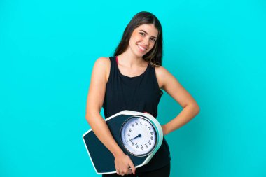 Genç İtalyan kadın mavi arka planda, kolları kalçasında ve tartı makinesiyle izole edilmiş.