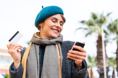 Açık havada esmer bir kadın kredi kartıyla cep telefonuyla alışveriş yapıyor.