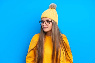 Mavi arka planda izole edilmiş kışlık giysiler giyen genç beyaz kadın yan tarafa bakıyor.