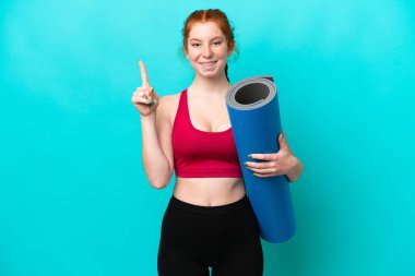 Young sport reddish woman going to yoga classes while holding a mat isolated on blue background pointing up a great idea