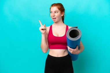 Genç spor kırmızısı kadın yoga derslerine giderken mavi arka planda izole edilmiş bir minder tutuyor. Çözümün farkına varmak için parmağını kaldırıyor.