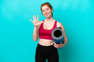Genç spor kırmızısı kadın yoga derslerine giderken elinde mavi arka planda izole edilmiş bir minder tutarken parmaklarıyla beşi sayıyor.